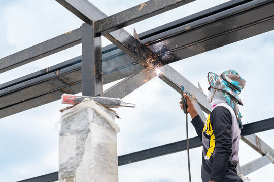 Construction worker welding a steel roof frame structure on a building site, generating bright sparks outdoors against the sky.