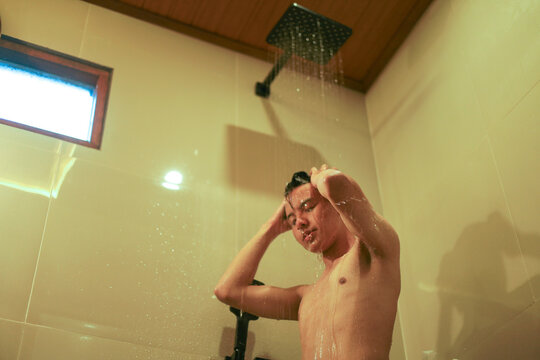 Young Asian Man Taking Refreshing Shower Under Modern Rain Showerhead in Warm Bathroom