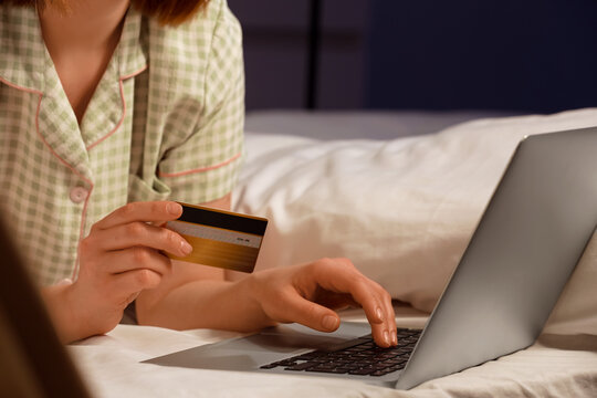 Young woman with credit card using laptop for online shopping on bed in evening at home, closeup