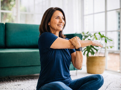 Cheerful middle-aged woman stretching her arm, sitting on a rug at home, promoting wellness.