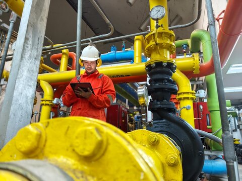 A low-angle shot of an industrial engineer in an orange jumpsuit, hardhat, and protective ear muffs holding a tablet while performing safety checks on a yellow gas pipeline system inside a facility.