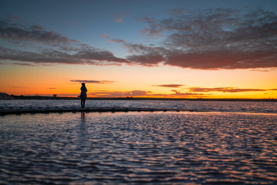A silhouetted woman figure standing at the end of a sweeping curved jetty, looking across calm waters toward a vivid sunset, Lake Tyrrell, Victoria, Australia
