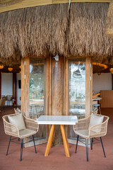 Fototapeta premium Empty cozy tropical cafe interior featuring a white table, woven rattan chairs with cushions, wooden flooring, and large wooden pillars against a white wall near glass folding doors.