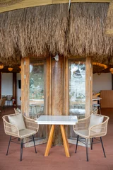 Fotobehang Horeca Empty cozy tropical cafe interior featuring a white table, woven rattan chairs with cushions, wooden flooring, and large wooden pillars against a white wall near glass folding doors.  © Yellow Boat