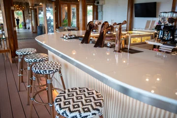 Fotobehang Horeca Modern tropical cafe interior featuring a pristine white counter, stylish patterned bar stools, and sleek metallic espresso taps under a rustic woven ceiling decorated with warm pendant lights.  © Yellow Boat