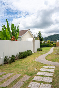 Modern white minimalist resort villa with an elegant archway entrance and solid wooden door, surrounded by tropical green plants and a white privacy wall under a bright blue cloudy sky.
