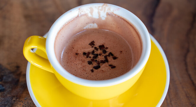 Partially consumed hot chocolate with cocoa nibs in a bright yellow ceramic cup and saucer resting on a rustic wooden table, offering a cozy and comforting cafe lifestyle experience.