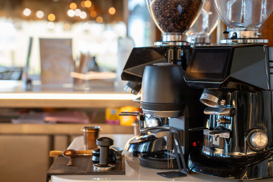 Modern black espresso coffee grinders displaying roasted beans in transparent hoppers on a cafe counter. Warm illuminated background highlighting professional barista equipment in a coffee shop.