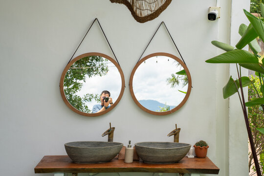 Twin round wooden mirrors hanging above natural stone washbasins on a solid wood counter against a white wall in a beautiful outdoor tropical resort bathroom setting with green reflections.