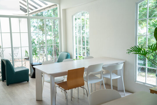 bright and modern cafe or restaurant interior featuring a large white dining table surrounded by minimalist chairs, with large windows overlooking a lush outdoor garden seating area.