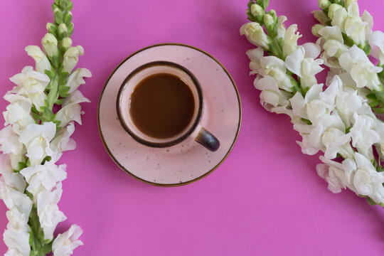 Soft pink background frames a warm cup of coffee. White flowers cascade gently on either side. Coffee cup sits centered, inviting and calm. Petals contrast with earthy tones, soft and elegant