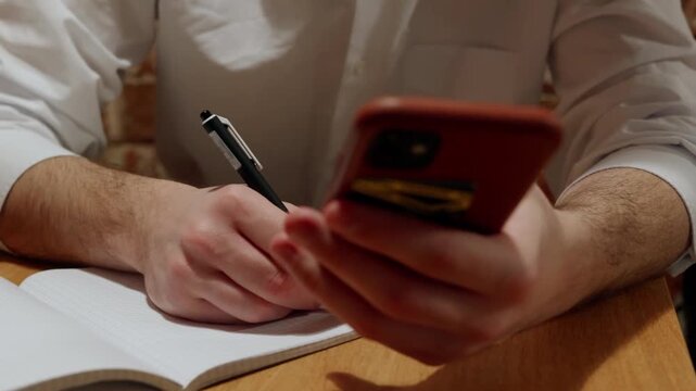 A person in white shirt writes in notebook while holding smartphone. Action progresses from poised pen to active writing as they reference device