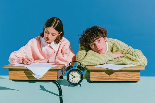 Black twin-bell alarm clock standing between two students at wooden desks, featuring knitted textures and a vivid blue background with copy space for school routine campaigns