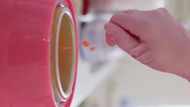 Vertical video. Wax grains. Skin care. Unrecognizable female hand in pink latex glove using orange product granules for melting in heater preparation for professional cosmetic procedure.