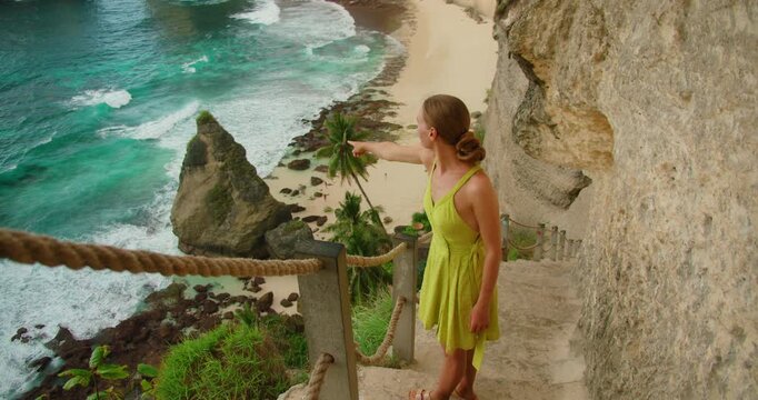 Woman pointing at tall cliff in turquoise ocean water from narrow trail leading to Diamond Beach in Bali