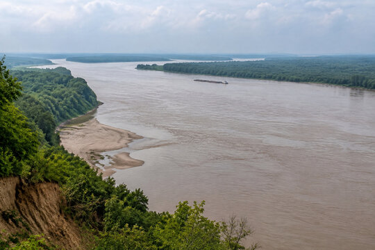 View from high bluff of wide brown river bending through low green forest, sandbar, tugboat and barge downstream, Mississippi River landscape