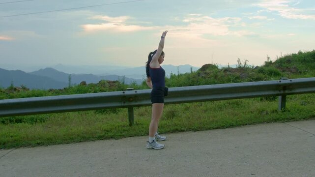 Young woman stretching and raising arm along a mountain road during sunset at Mau Son, Vietnam. Outdoor fitness, cardio recovery, and active travel lifestyle