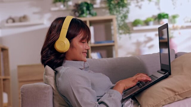 Woman Wearing Headphone Using Laptop On Sofa In Living Room At Home Smiling During Remote Work Online Communication Typing Learning Screen Relaxed Technology Comfort Internet Digital Audio