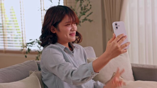 Asian woman taking smartphone selfie with smile on sofa at home in cozy living room with curtain window pillow plant warm indoor light casual peace sign video call