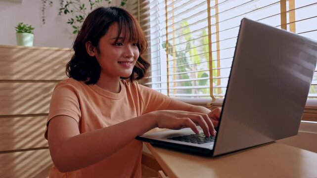 Asian woman typing on laptop at home office desk near window with plant, casual indoor workspace, warm light, focused smile, remote work setup