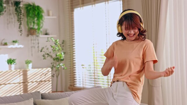 Young woman headphone music smile dance living room casual home lifestyle scene with plant window curtain joyful expression bright interior warm sunlight green decor