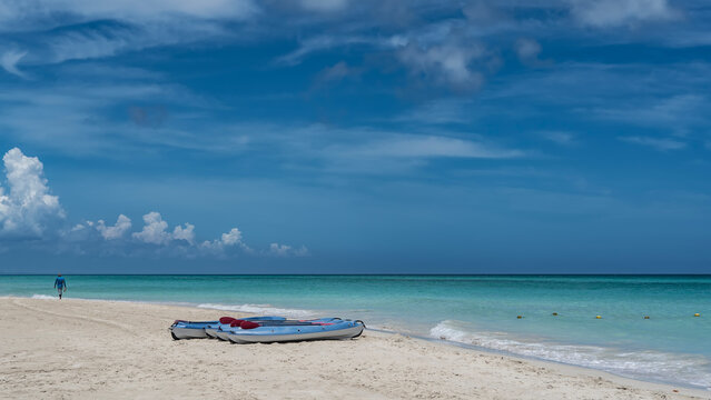 Tourist canoes with oars on a sandy beach. The silhouette of a person walking along the seashore. The waves of the aquamarine ocean foam on the white sand. Blue sky, clouds. Cuba. Varadero.