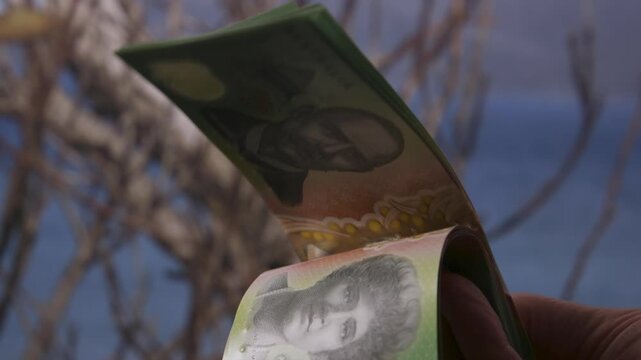 Human fingers flip through a curved stack of Australian 100 dollar banknotes against an outdoor background.