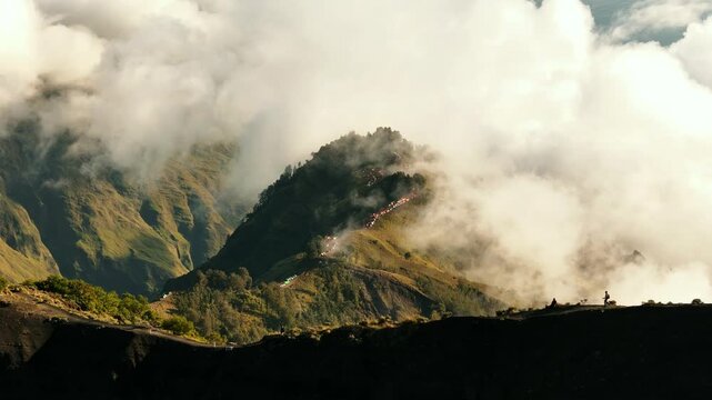 Ethereal clouds drift across Mount Rinjani ridges above lush volcanic slopes in golden morning light, aerial pullback