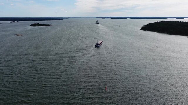 Small general cargo ship navigating through Finnish archipelago. Vessel passing narrow fairway close to shoreline. Ascending aerial stern view revealing the passage ahead.