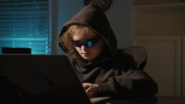 Night hacker child working on computer. Hooded hacker kid in cyber crime activity. Hacker child attack on digital system. Hacker kid typing code in laptop at night office.