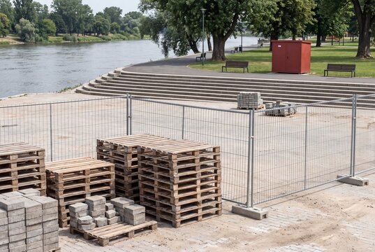 Construction site near a river features stacks of wooden pallets and paving stones enclosed by a metal fence.