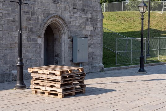 Stack of wooden pallets sits on a paved walkway outside a stone building with an arched doorway