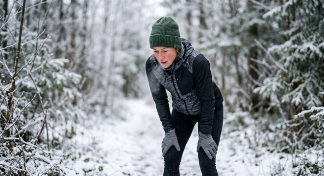 A weary woman wearing a green beanie and dark athletic wear taking a break to catch her breath during a run on a snowy forest trail in winter.
