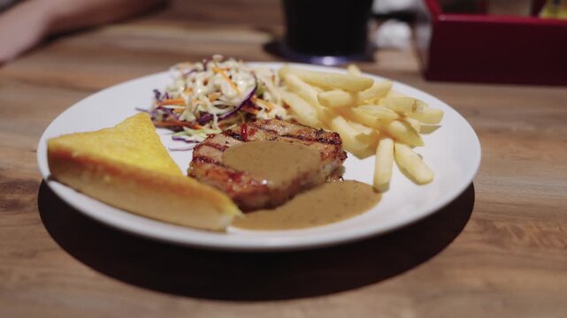 Grilled pork steak with black pepper gravy served with crispy french fries, coleslaw, and toasted buttered bread on a white plate. Close-up of a delicious restaurant meal on a wooden table.
