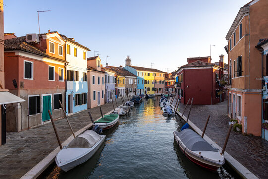 Colorful houses and moored boats line a quiet, empty canal in Burano at sunset, Venice, Italy