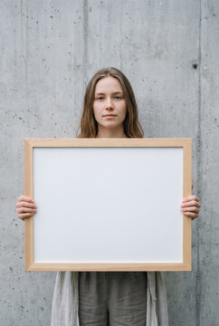 Young woman holding empty wooden picture frame against concrete wall portrait