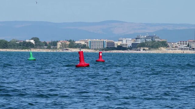 Red and green navigational buoys bob in the deep blue sea as seagulls fly past, with a wide sandy beach and a coastal resort town visible in the distance under a clear sky.