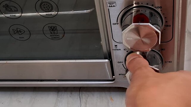 A close up view shows a hand turning a dial on a kitchen oven preparing to cook food with illuminated heating elements
