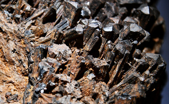 A macro close-up view of an ilvaite mineral specimen featuring well-developed, dark metallic black prismatic crystals growing in a cluster on a brownish rock matrix.
