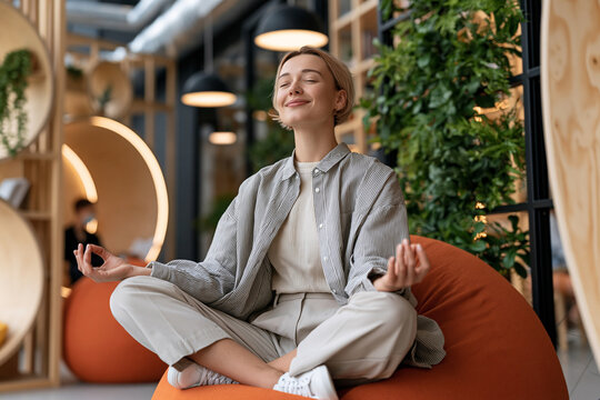 Woman sits calmly in a modern, plant-filled space. Her closed eyes and serene expression suggest deep peace. Fingers form a gentle mudra, signaling mindfulness