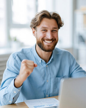 Bearded man smiles broadly, fists clenched in excitement. He sits at a desk, laptop open beside papers. Bright window light illuminates his joyful expression