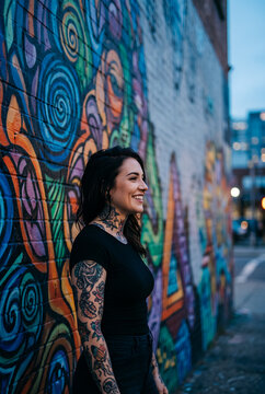 Young woman with tattoo smiling at colorful street graffiti wall wearing black