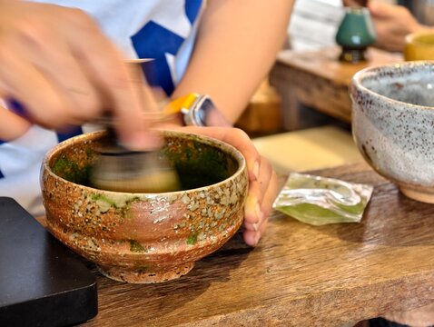 Action shot of a person whisking Japanese matcha green tea in a traditional ceramic bowl with a bamboo whisk. Motion blur effect showing authentic tea preparation and cafe beverage concepts.