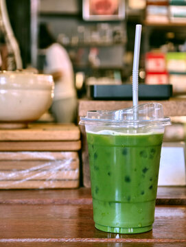 Close-up of refreshing iced Japanese matcha green tea in a clear plastic cup with straw. Placed on a wooden table with blurred cafe background. Perfect for beverage menu and coffee shop concepts.