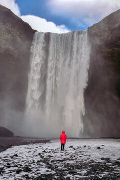 Majestic Skogafoss waterfall in Iceland with a traveler in a red jacket standing in awe of nature&rsquo;s raw power and beauty.