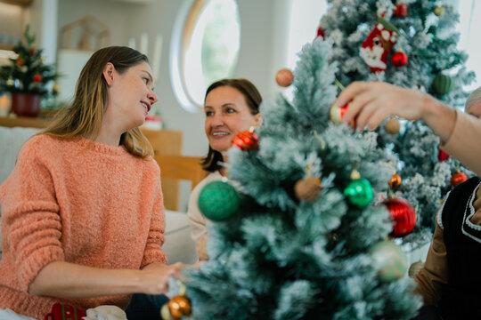 Adult daughter and senior parents decorating Christmas tree together at home, sharing conversation, happiness, family bonding, festive holiday tradition.