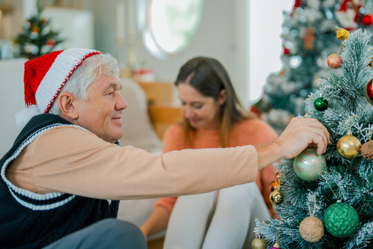 Senior father wearing Santa hat decorating Christmas tree with ornaments while spending festive holiday time with his family at home.