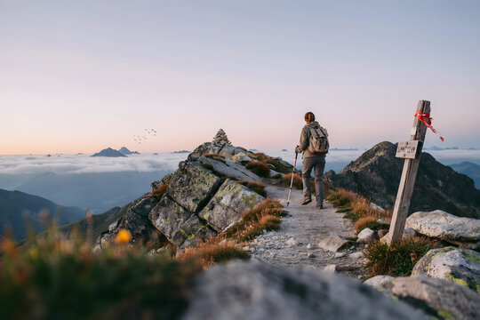A hiker with a backpack and trekking pole walking on a rocky mountain ridge trail towards a stone cairn at sunset with a wooden trail marker