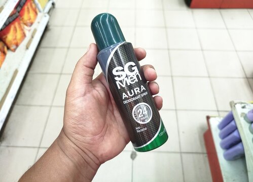 KUALA TERENGGANU, MALAYSIA- AUGUST 20. 2026: Close-up of a hand holding SG Men Aura deodorant spray bottle inside a hypermarket aisle, with tiled floor and product shelves in the background.