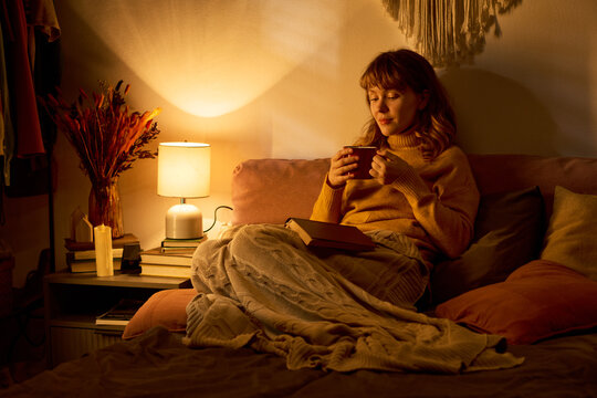 Caucasian young adult woman sitting in bed holding mug, relaxing during evening leisure time. Image suitable for mental wellbeing campaigns and home lifestyle marketing